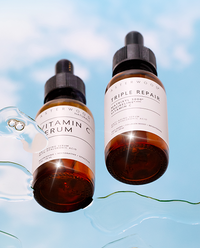 Asterwood Vitamin C Serum and Triple Repair serum bottles floating against a sky-blue cloud backdrop with water trails.
