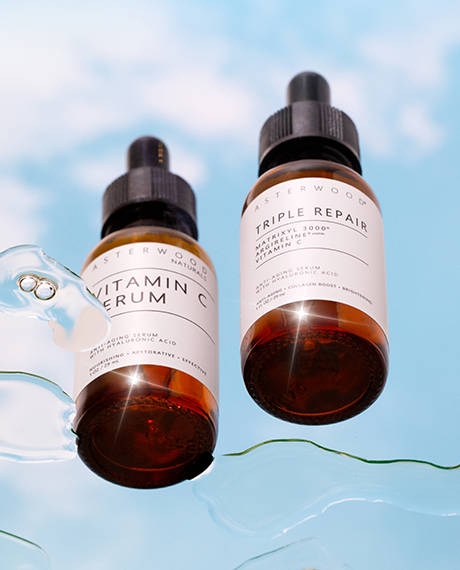Asterwood Vitamin C Serum and Triple Repair serum bottles floating against a sky-blue cloud backdrop with water trails.