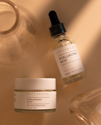 Top-down Asterwood Hyaluronic Acid serum and moisturizer jar arranged with glass props on a warm tan surface with sunlit shadows.