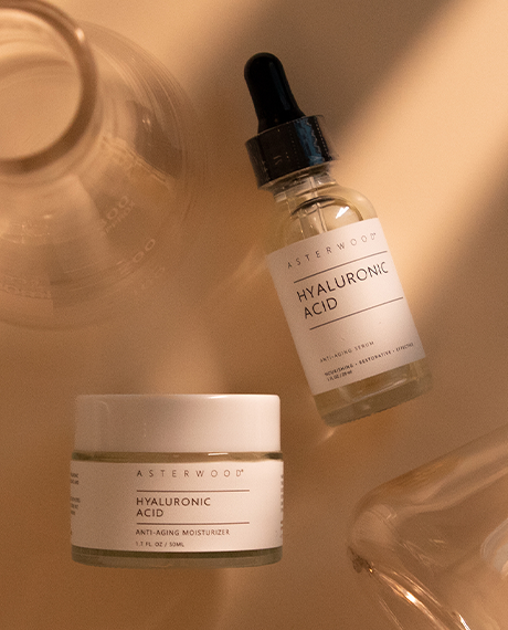 Top-down Asterwood Hyaluronic Acid serum and moisturizer jar arranged with glass props on a warm tan surface with sunlit shadows.