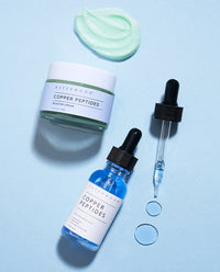 Flat lay on blue of the Copper Peptides Booster Cream jar and blue Copper Peptides Serum with its dropper, mint cream swatch and blue serum drops.