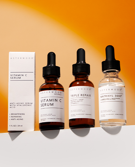 Asterwood Powerhouse Trio lined up on a warm orange background: the Vitamin C Serum carton with the amber Vitamin C Serum, amber Triple Repair Serum, and clear Matrixyl 3000 + Argireline Serum bottles.