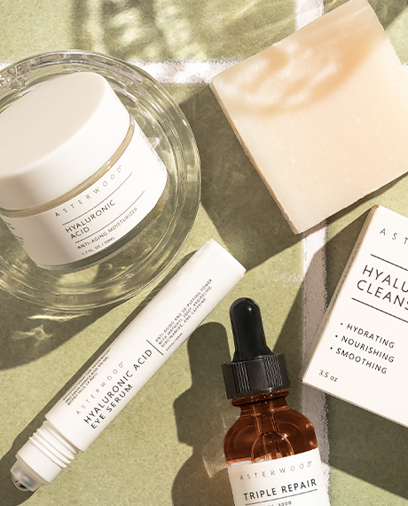 Top-down lifestyle of the Skin Rescue Routine: HA Moisturizer, HA Cleansing Soap bar with carton, HA Eye Serum pen, Triple Repair on green stone.