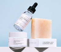 Hyaluronic Acid Cleansing Soap bar perched with HA serum bottle above and HA Moisturizer jar at left against a soft blue background.