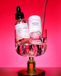 Asterwood Hyaluronic Acid serum and Volulip Plumping Lip Treatment perched in a cocktail glass with ice against a vibrant pink background.