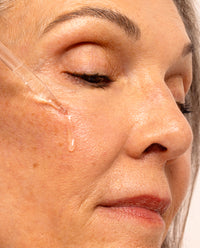 Mature woman with grey hair applying Asterwood Argireline Serum to her cheek with the glass dropper, a drop visible against her skin.