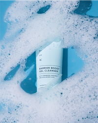 Asterwood Barrier Boost Gel Cleanser tube partially submerged in white cleansing foam on a blue background.