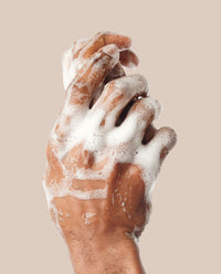 Hands working a thick white lather from the Hyaluronic Acid Cleansing Soap against a tan background.