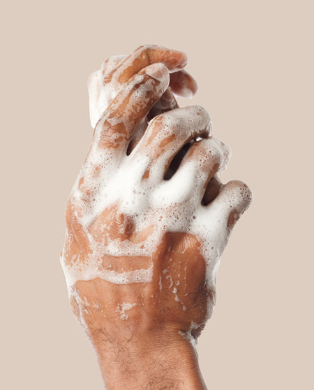 Hands working a thick white lather from the Hyaluronic Acid Cleansing Soap against a tan background.
