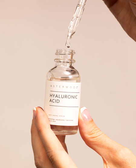Hand holding the clear Asterwood Hyaluronic Acid Serum with the dropper releasing a droplet above the open bottle.
