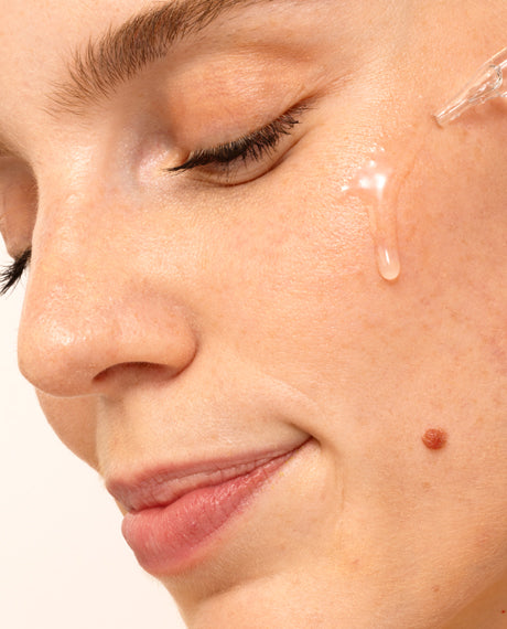 Close-up of a woman's cheek with a clear droplet of Matrixyl 3000 + Argireline serum tracing down her skin from a glass dropper.
