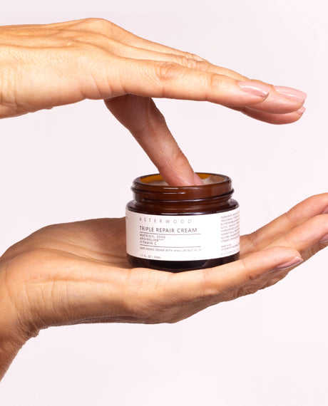 Hand dipping a finger into the open Asterwood Triple Repair Cream amber jar, scooping the ivory anti-aging cream.