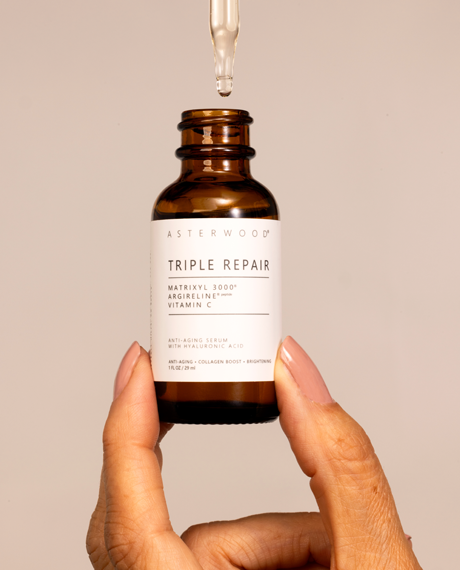 Hand holding Asterwood Triple Repair amber serum with dropper releasing a clear bead onto the label, beige background.