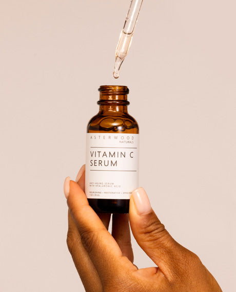 Hand holding the Asterwood Vitamin C Serum amber bottle with a glass dropper above releasing a golden bead onto the open bottle.