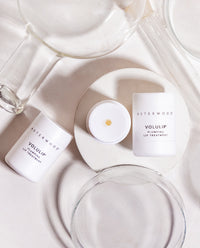 Flat lay of two Asterwood Volulip Plumping Lip Treatment tubes among clear glass dishes, one open showing a golden balm dot.