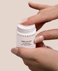 Hands holding the open Asterwood Volulip Plumping Lip Treatment, a glossy bead of balm at the twist-up base.